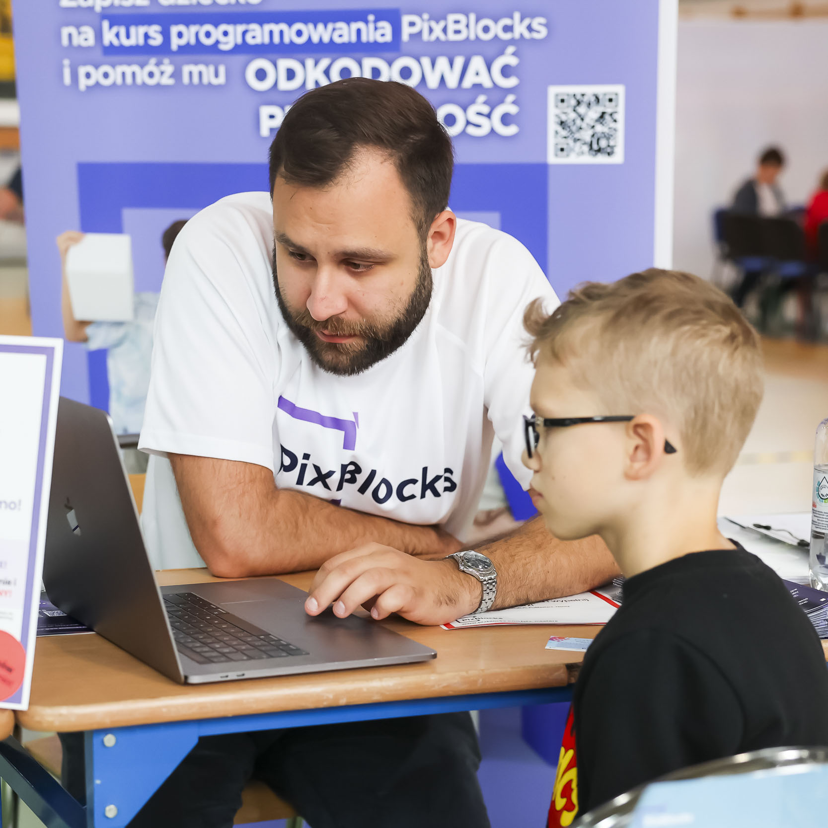 Opiekun warsztatów pokazujący dziecku treści w laptopie, za nimi ścianka kursu programowania PixBlocks