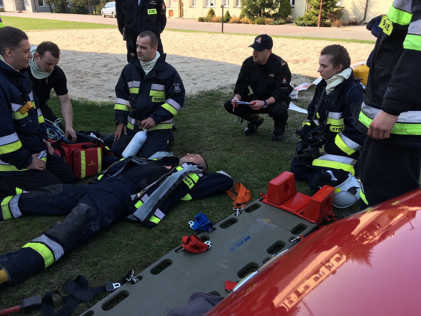 Zdjęcie przedstawia grupę strażaków OSP, instruktora PSP oraz pozoranta leżącego na ziemi w trakcie omawiania ćwiczenia.