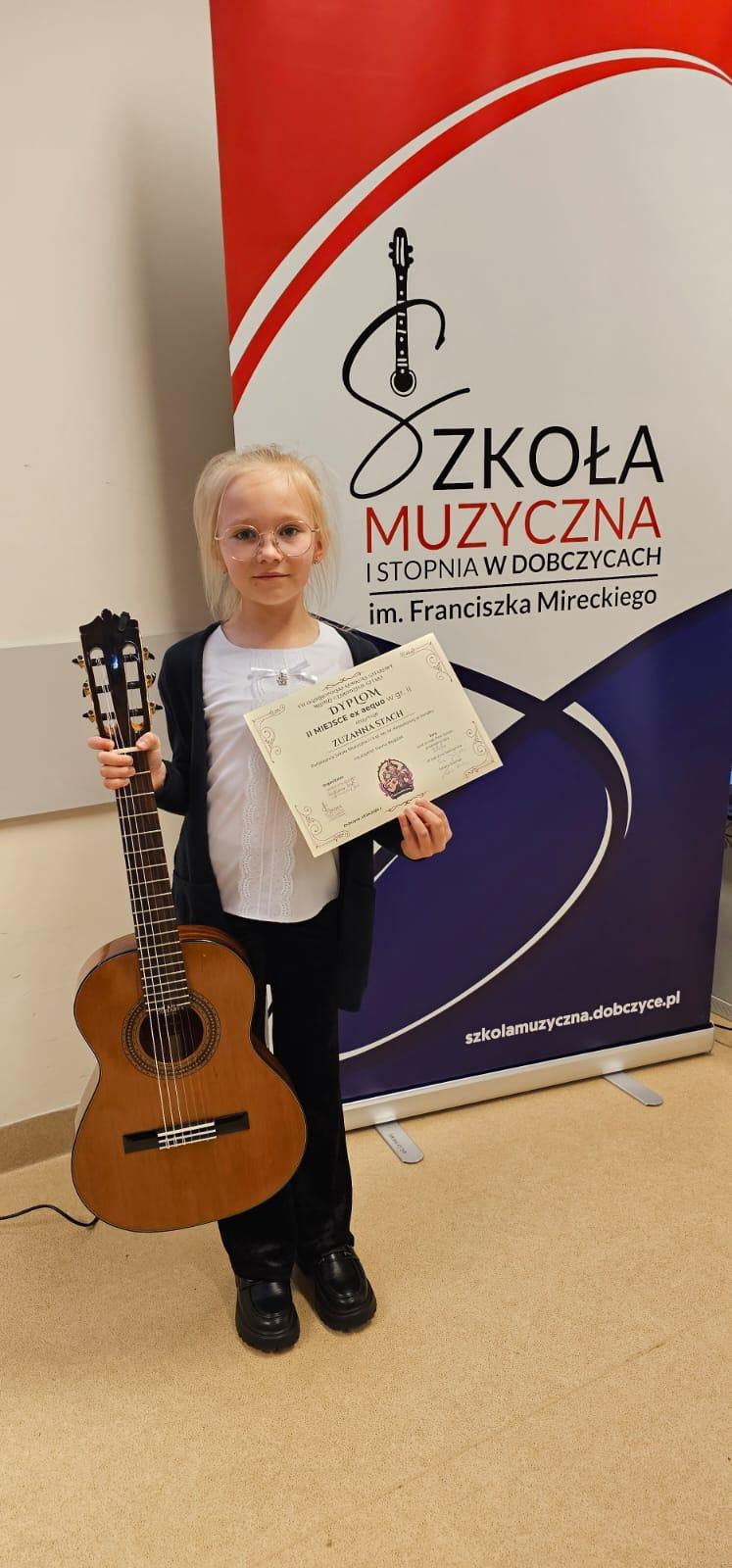 Zuzanna Stach w VII Ogólnopolskim Konkursie Gitarowym „Młodzi czarodzieje gitary” w Dobczycach. Zdjęcie uczennicy z gitarą, w tle rollup reklamujący konkurs.