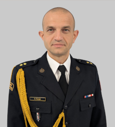 Mężczyzna w mundurze galowym stoi na neutralnym, jasnoszarym tle. Ma krótkie, ogolone włosy i patrzy prosto w obiektyw z poważnym wyrazem twarzy. Ubrany jest w ciemnogranatowy mundur z pagonami ozdobionymi w stopniu kapitana oraz złotym sznurem naramiennym na prawym ramieniu. Na lewej piersi widoczna jest plakietka z nazwiskiem ‘D. PAJĄK’ oraz dwie wstążki odznaczeń. Mundur posiada również emblematy i naszywki wskazujące na służbę mundurową