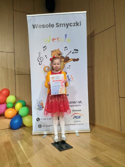 Uczennica w czerwonej sukience i żółtym sweterku trzymająca dyplom wyróżnienia. Po lewej stronie widoczne kolorowe balony.