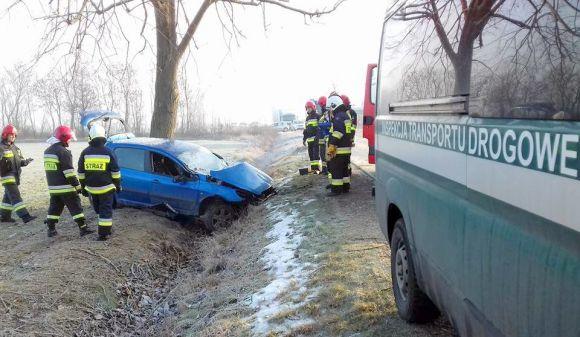 Na pierwszym planie samochód ITD, dalej strażacy stojący wokół samochodu osobowego z rozbitym przodem i częściowo w rowie