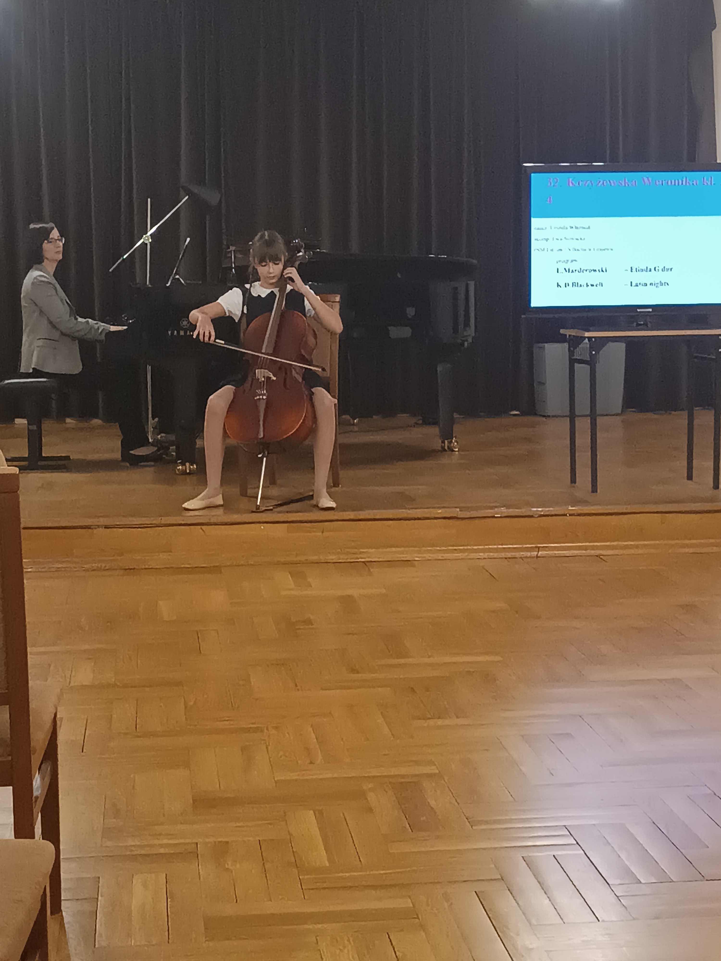 Zdjęcie przedstawia Weronikę Krzyżewską grającą na wiolonczeli podczas zmagań konkursowych. W tle pani Ewa Nowacka akompaniująca uczennicy. Po prawej stronie widać ekran, na którym wyświetlone są informacje o repertuarze jaki wykonuje Weronika Krzyżewska. 