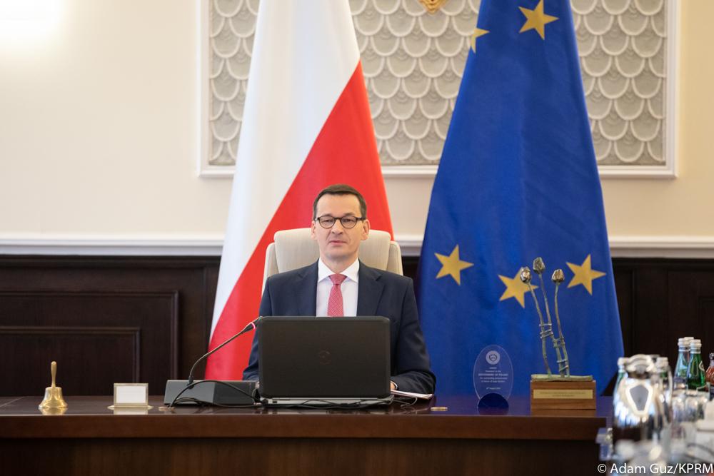 Premier Mateusz Morawiecki siedzi na tle flagi Polski i flagi UE.