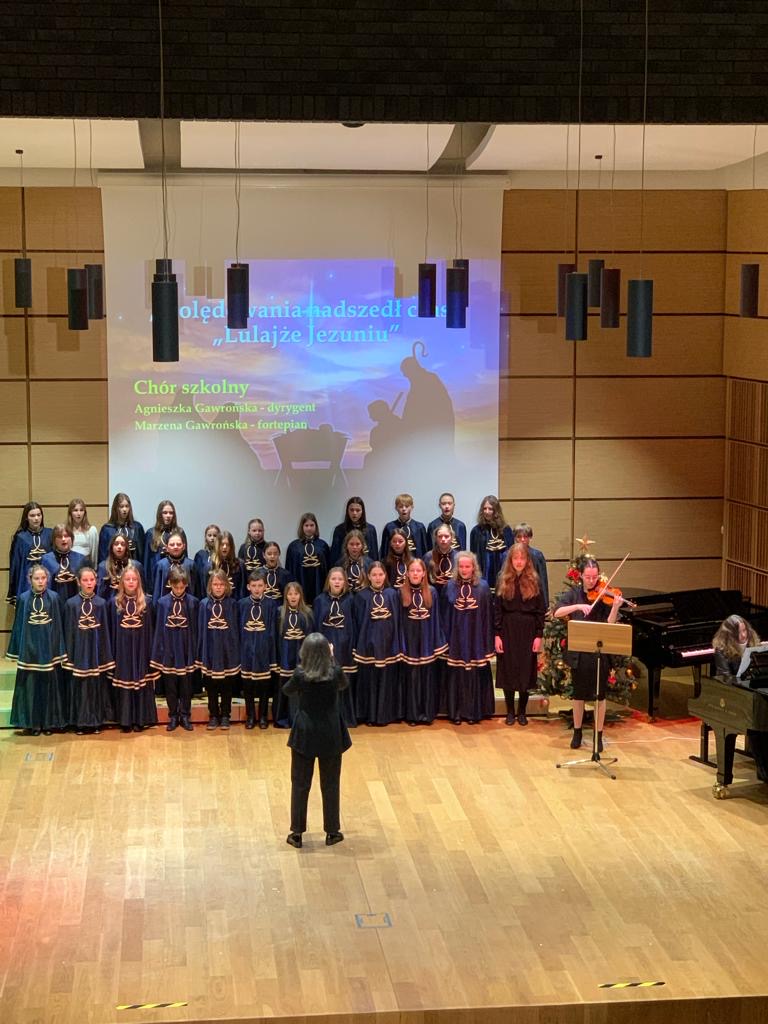 Chór PSM w Tarnobrzegu śpiewający z towarzyszeniem skrzypiec oraz fortepianu. Przed chórem stoi Pani Dyrygent.