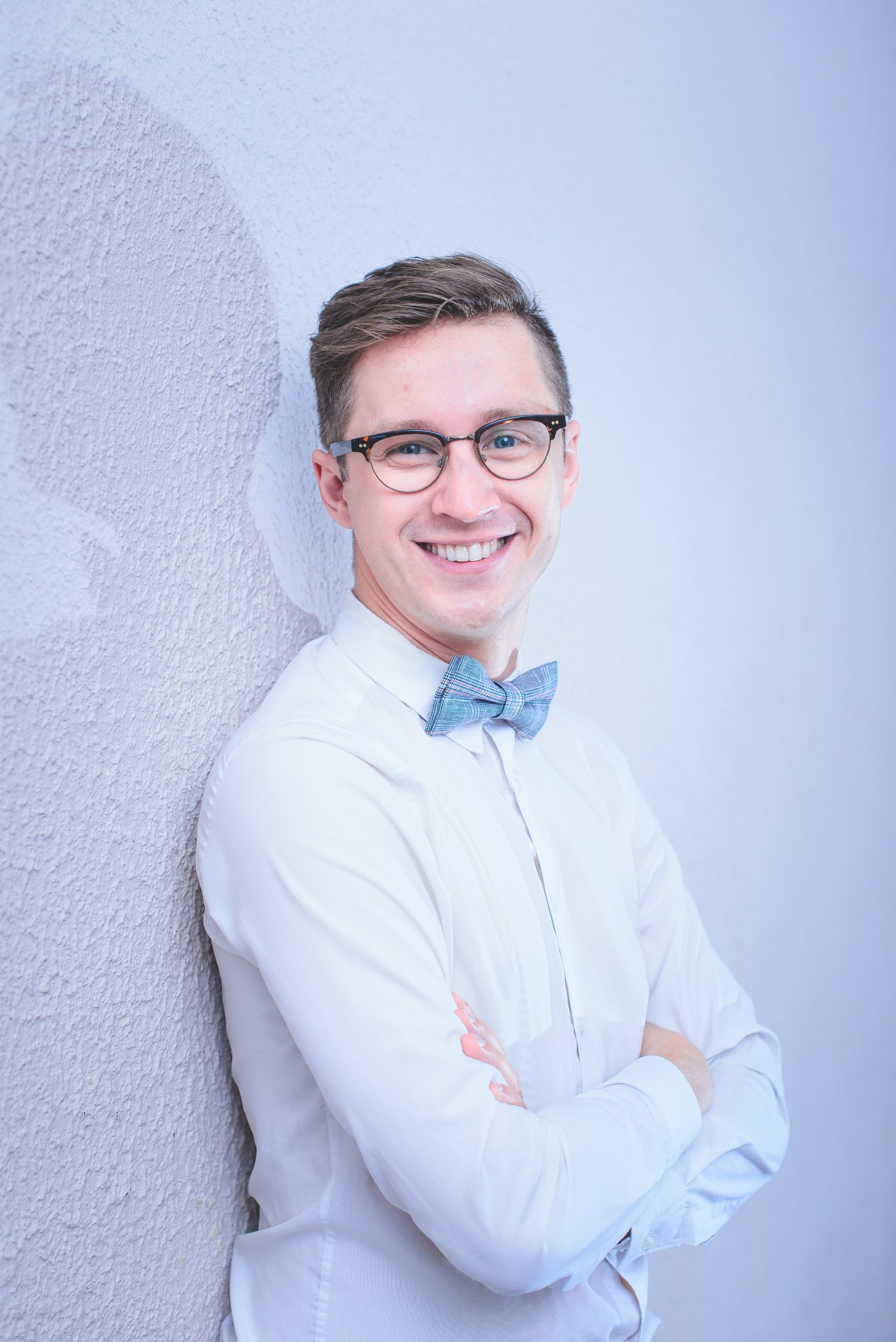 Mężczyzna ubrany w białą koszulę stoi oparty o białą ścianę. Mężczyzna uśmiecha się, ma okulary i niebieską muszkę.