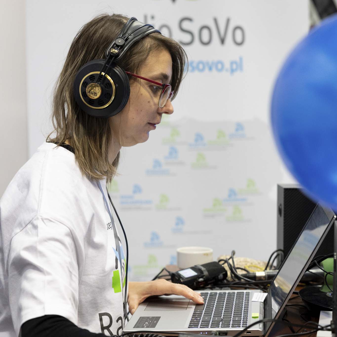 Młoda dziewczyna siedzi przed laptopem. Ma założone słuchawki, pracuje na laptopie.