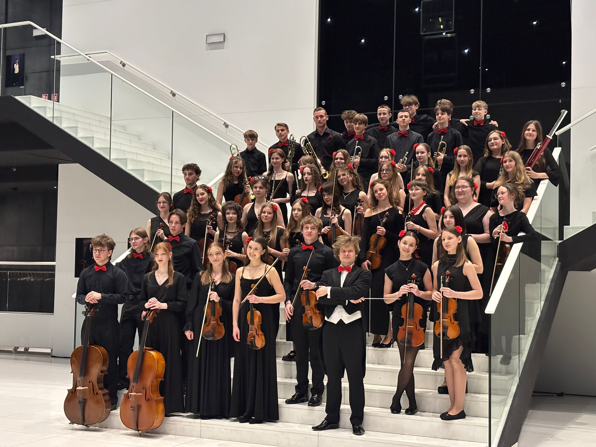 Zdjęcie grupowe młodzieżowej orkiestry symfonicznej, wykonane w nowoczesnym, jasnym wnętrzu na tle białych schodów z szklaną balustradą. Muzycy, ubrani w eleganckie czarne stroje wizytowe z czerwonymi akcentami – chłopcy w czerwonych muchach, a dziewczęta z czerwonymi kwiatami we włosach – pozują w kilku rzędach, trzymając swoje instrumenty. W pierwszym rzędzie widoczne są dwie wiolonczele, liczne skrzypce oraz dyrygent w czarnym fraku z białą kamizelką i czerwoną muchą, stojący dumnie w centrum z batutą. W wyższych rzędach na schodach ustawieni są pozostali członkowie zespołu, w tym sekcja dęta z trąbkami, puzonami i fletami oraz fagocistka. Całość kompozycji emanuje profesjonalizmem i uroczystą atmosferą po występie, a duże przeszklenia w tle odbijają punktowe światła sufitowe, dodając zdjęciu głębi.