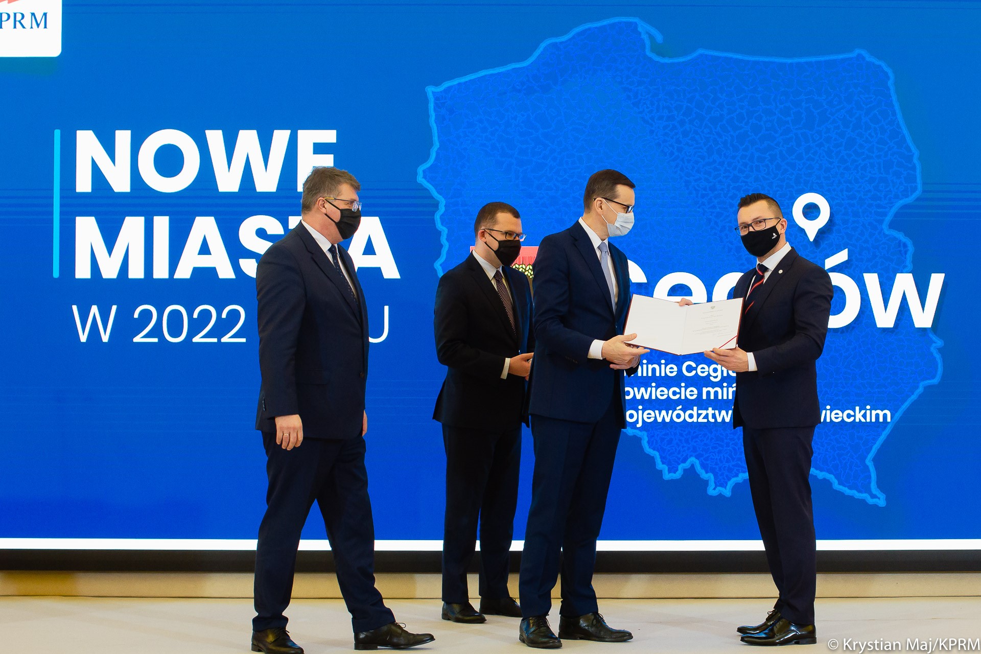 Premier wręcza akt nadania praw miejskich, obok stoi minister Paweł Szefernaker i minister Maciej Wąsik.