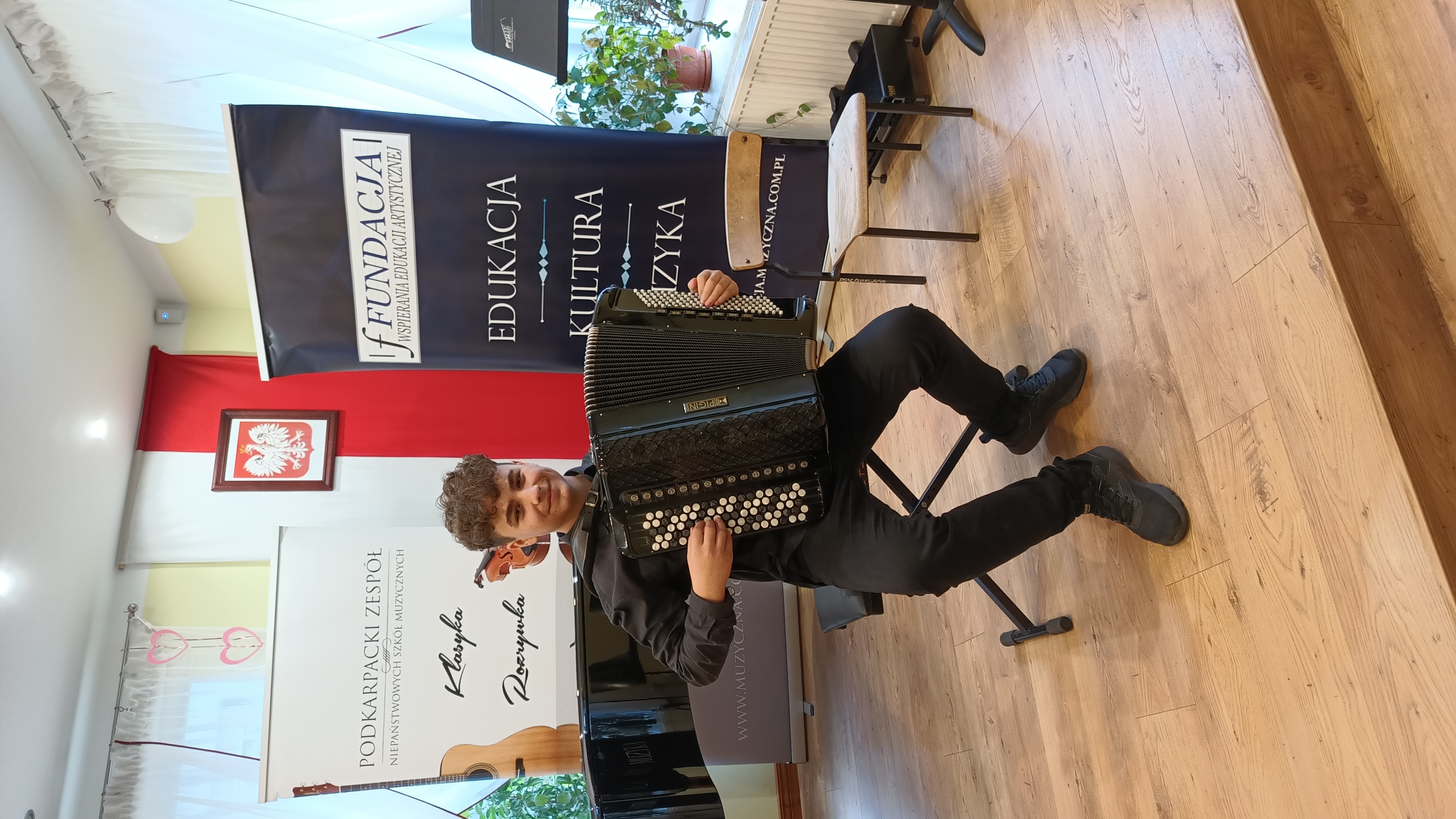 Na zdjęciu uśmiechnięty uczeń siedzący z akordeonem, w tle baner organizatorów.