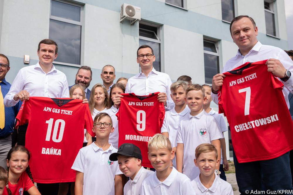 Premier Mateusz Morawiecki, minister Witold Bańka i marszałek JakubChełstowski stoją wśród dzieci trzymając koszulki z numerami i swoimi nazwiskami.