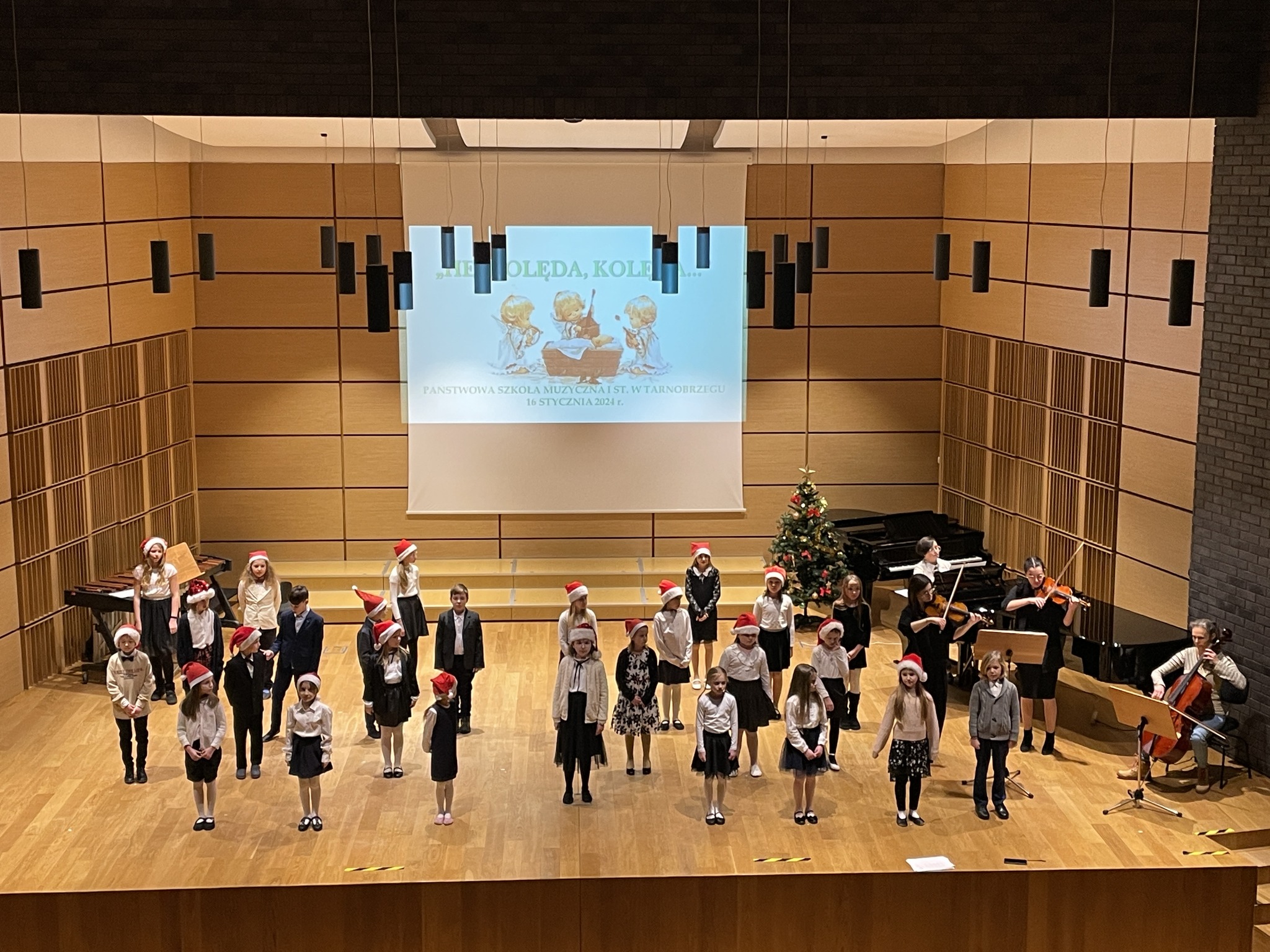 Śpiewający uczniowie wraz z grającymi na fortepianie, skrzypcach i wiolonczeli nauczycielami na scenie.