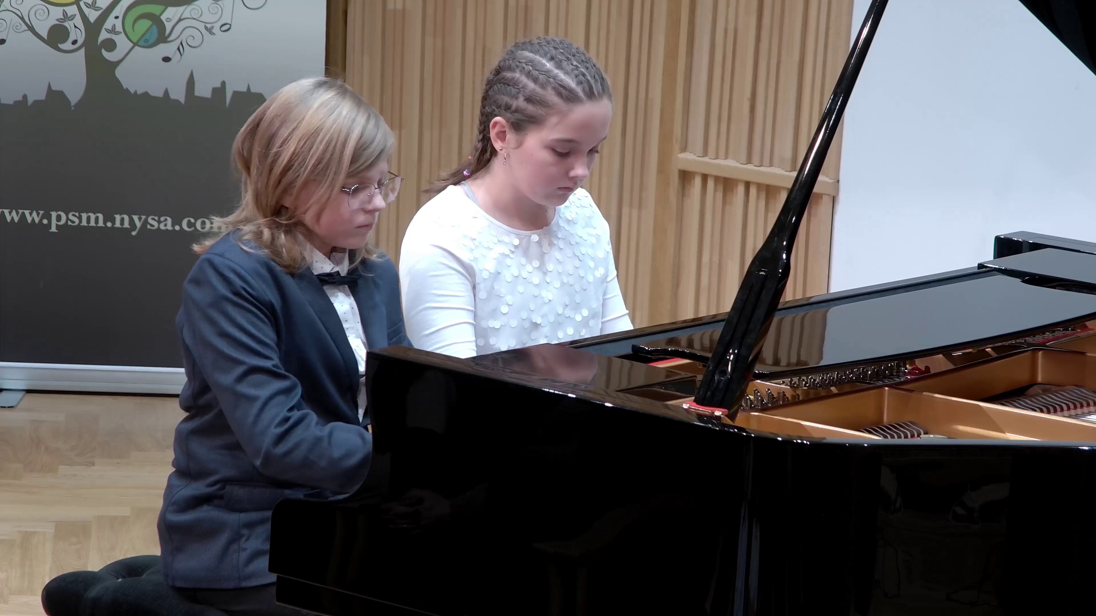 Zdjęcie. Estrada Sali Koncertowej. Na dużym czarnym fortepianie gra duet fortepianowy, z lewej uczeń, z prawej uczennica.