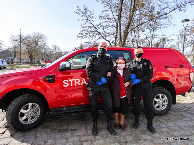 Na zdjęciu widać strażacki samochód. Na jego tle dwóch strażaków i Seniorka, tuż po szczepieniu na COVID-19