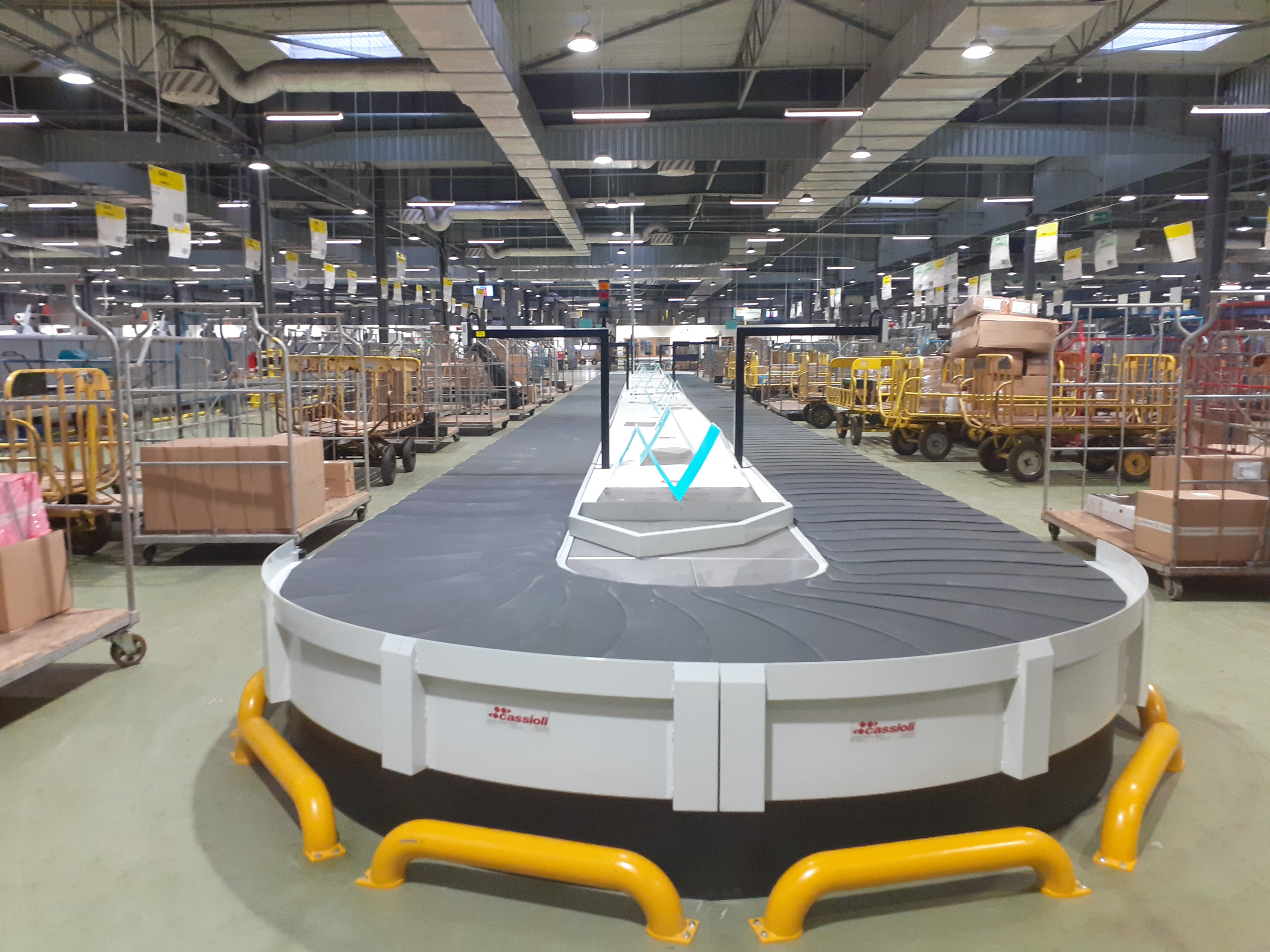 Hala magazynowa. Na środku taśma do sortowania, wokół niej żółte, niskie barierki. W całej hali stoi wiele wózków z paczkami