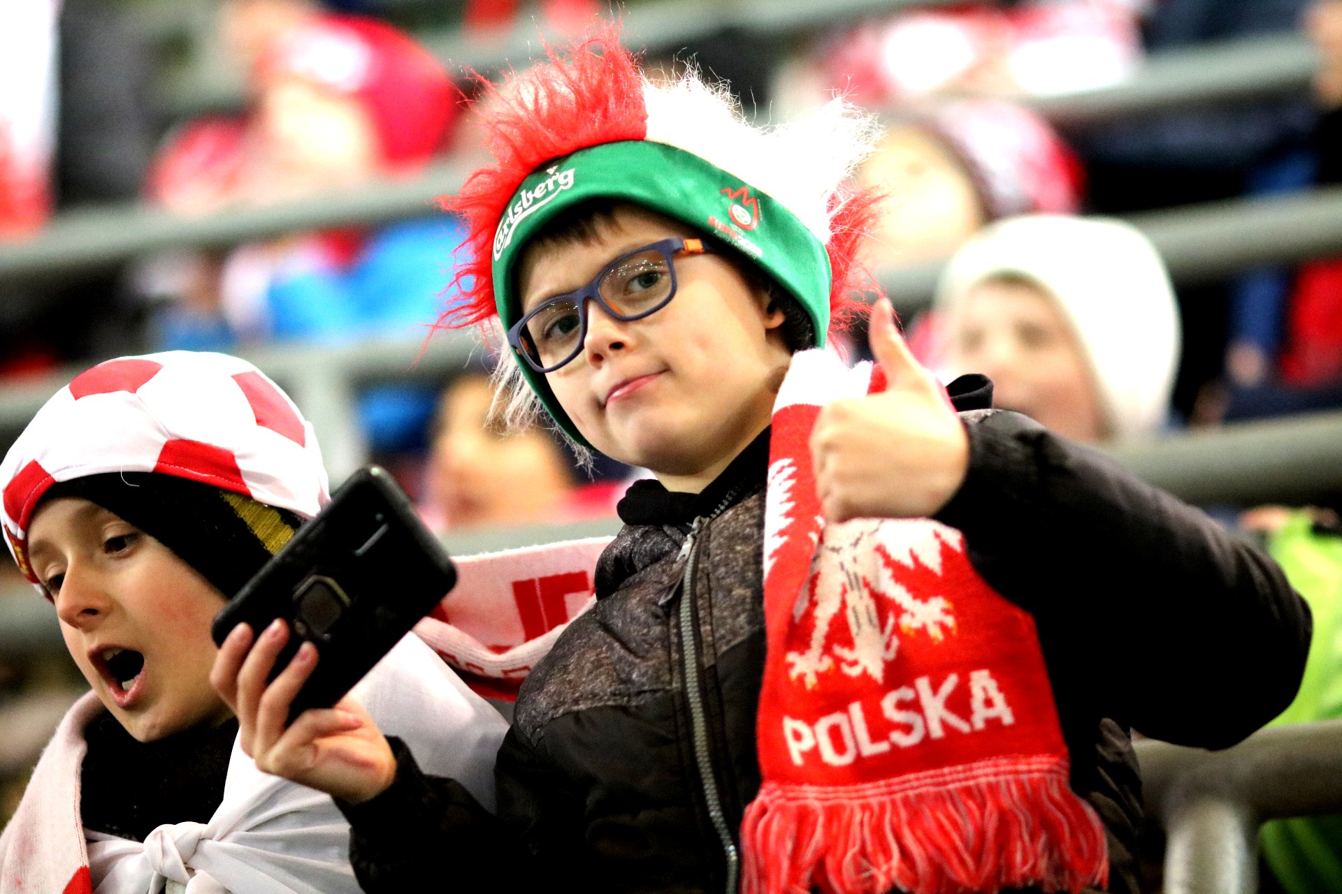 Chłopiec w okularach, czarnej kurtce, puchatej biało-czerwonej czapce z zielonym brzegiem i biało-czerwonym szaliku stoi na trybunie stadionu, w jednej ręce trzyma telefon a drugą pokazuje kciuk w górę.