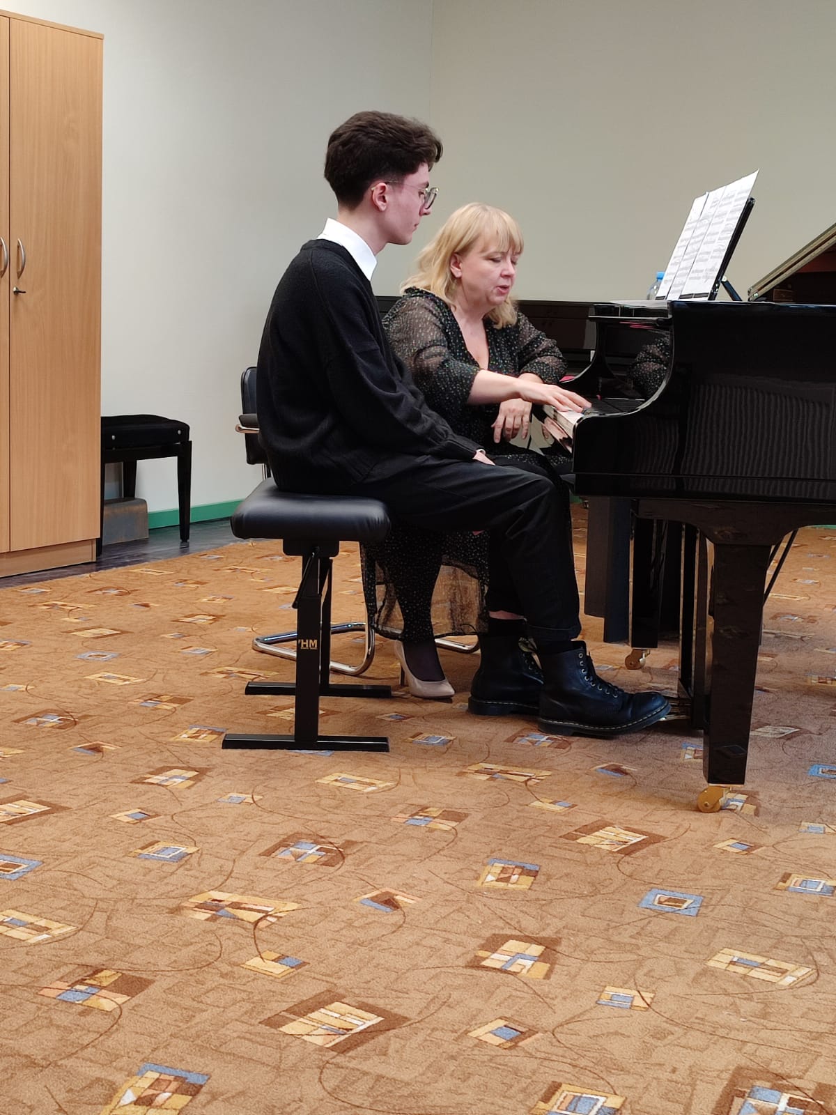 Uczeń siedzący przy fortepianie, obok niego nauczycielka. Po lewej stronie widoczna szafa.