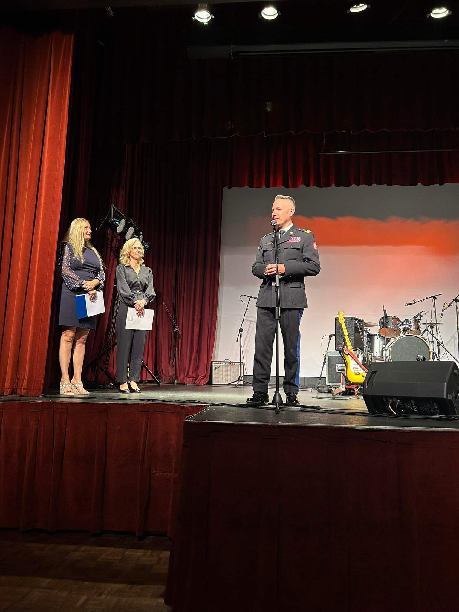 Komendant główny PSP ubrany w mundur galowy przemawia do mikrofonu na scenie, obok stoją dwie kobiety w ciemnych ubraniach