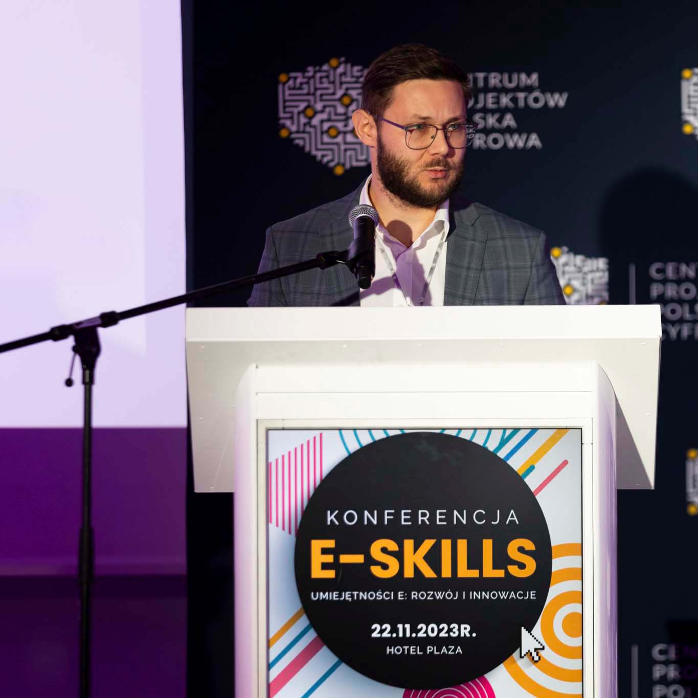 Przy mównicy stoi Pan Dawid Hoinkis. W ręku trzyma mikrofon patrząc na siedzących przed nią uczestników konferencji.