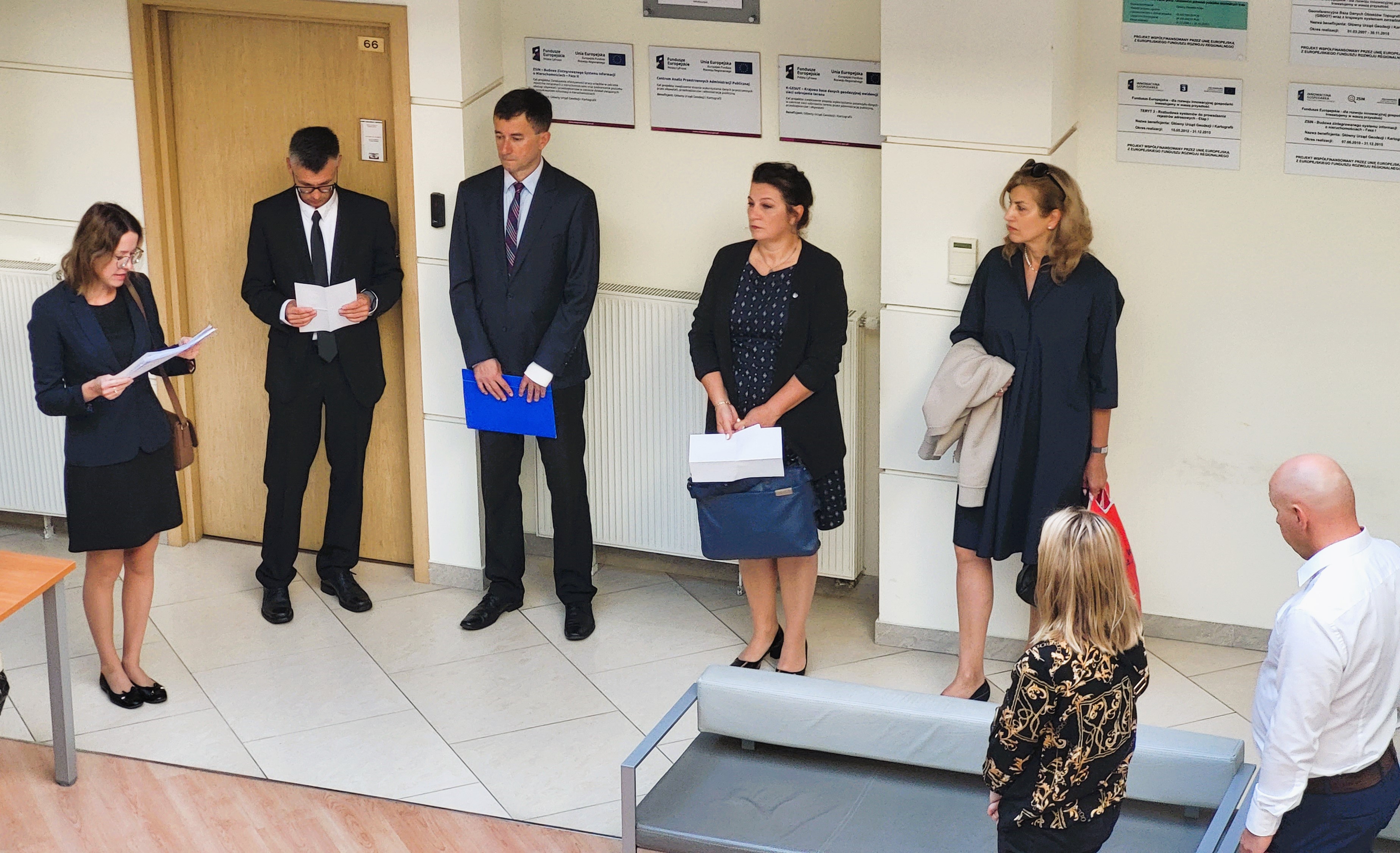 Zdjęcie przedstawia hol w siedzibie GUGiK przy ul. Olbrachta 94B z osobami przystępującymi do egzaminu słuchającymi wyjaśnień członków Komisji Egzaminacyjnej.