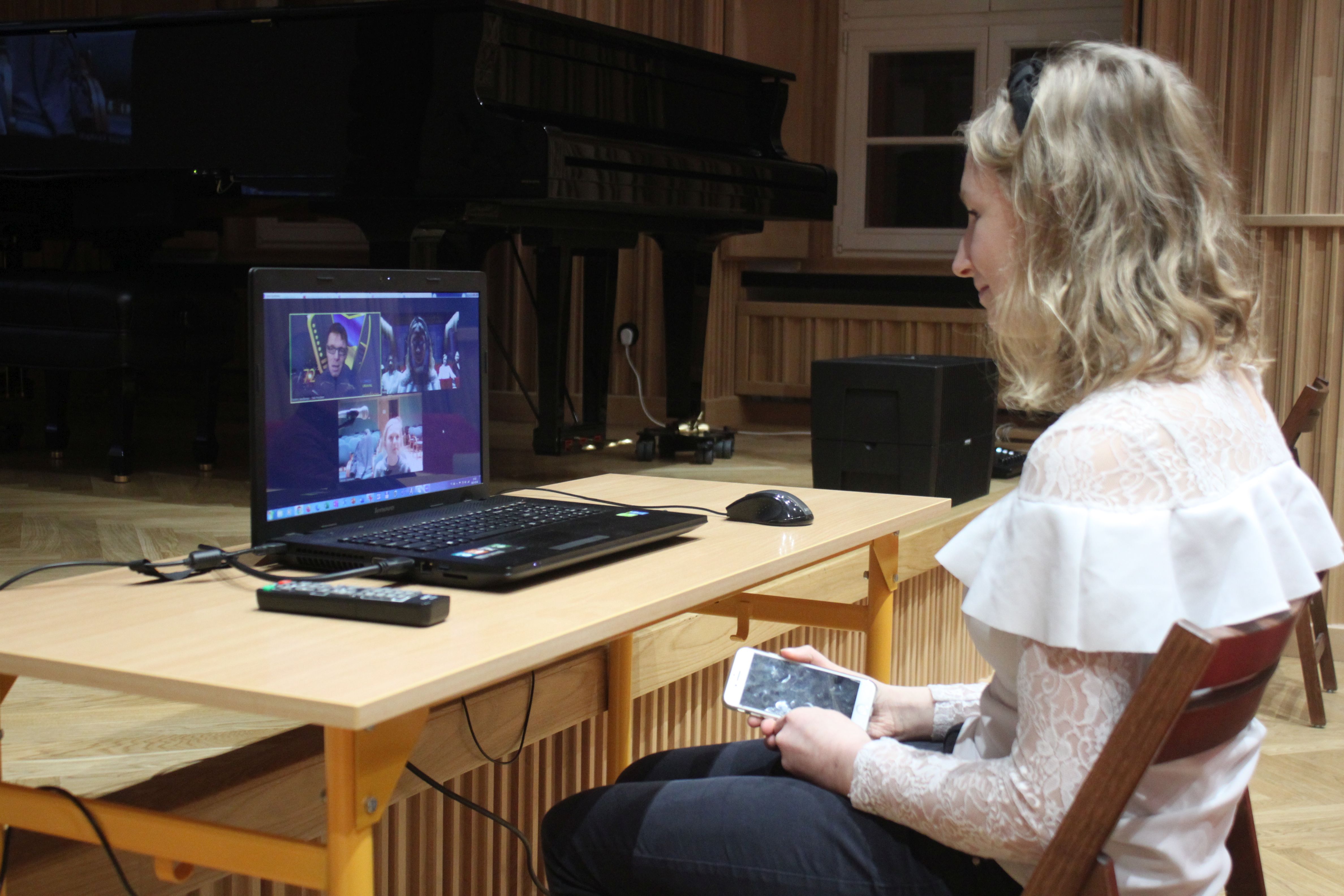 Zdjęcie. W Sali Koncertowej PSM w Nysie, przy ławce, na której stoi otwarty laptop, siedzi uczennica, która zadaje pytanie w spotkaniu online.
