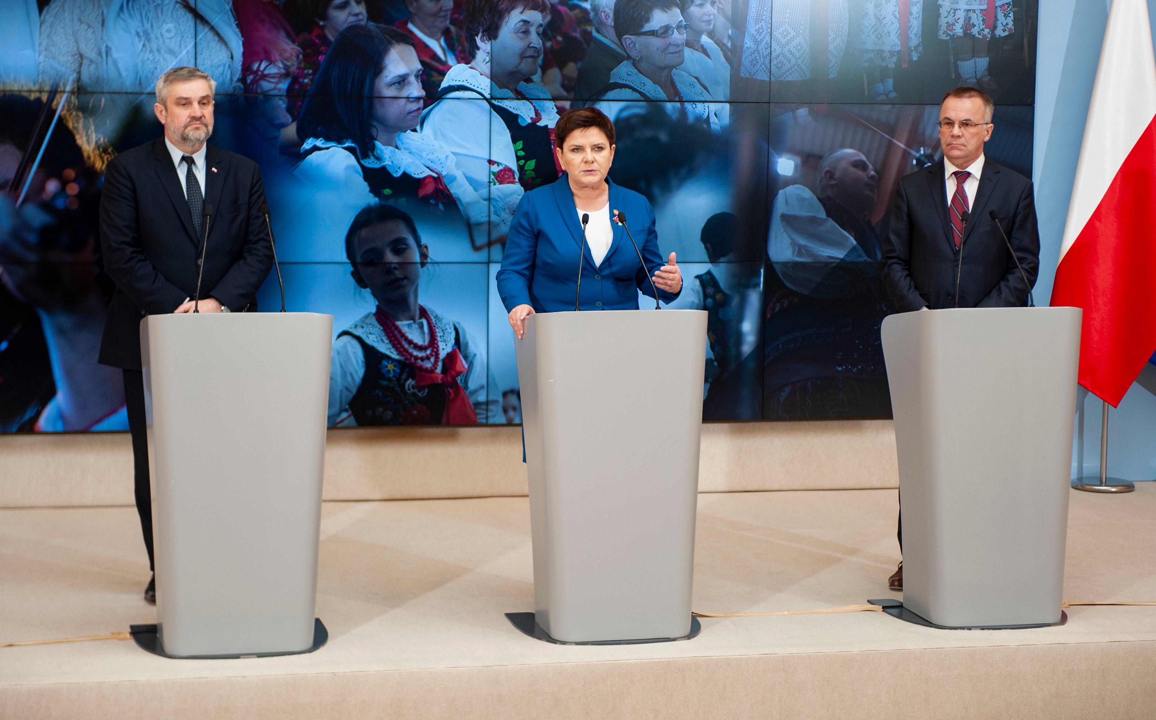Minister Jan Krzysztof Ardanowski, wicepremier Beata Szydło i minister Jarosław Sellin stoją przy mównicach, a za nimi ekran na którym wyświetlone jest zdjęcie kobiet w strojach ludowych.