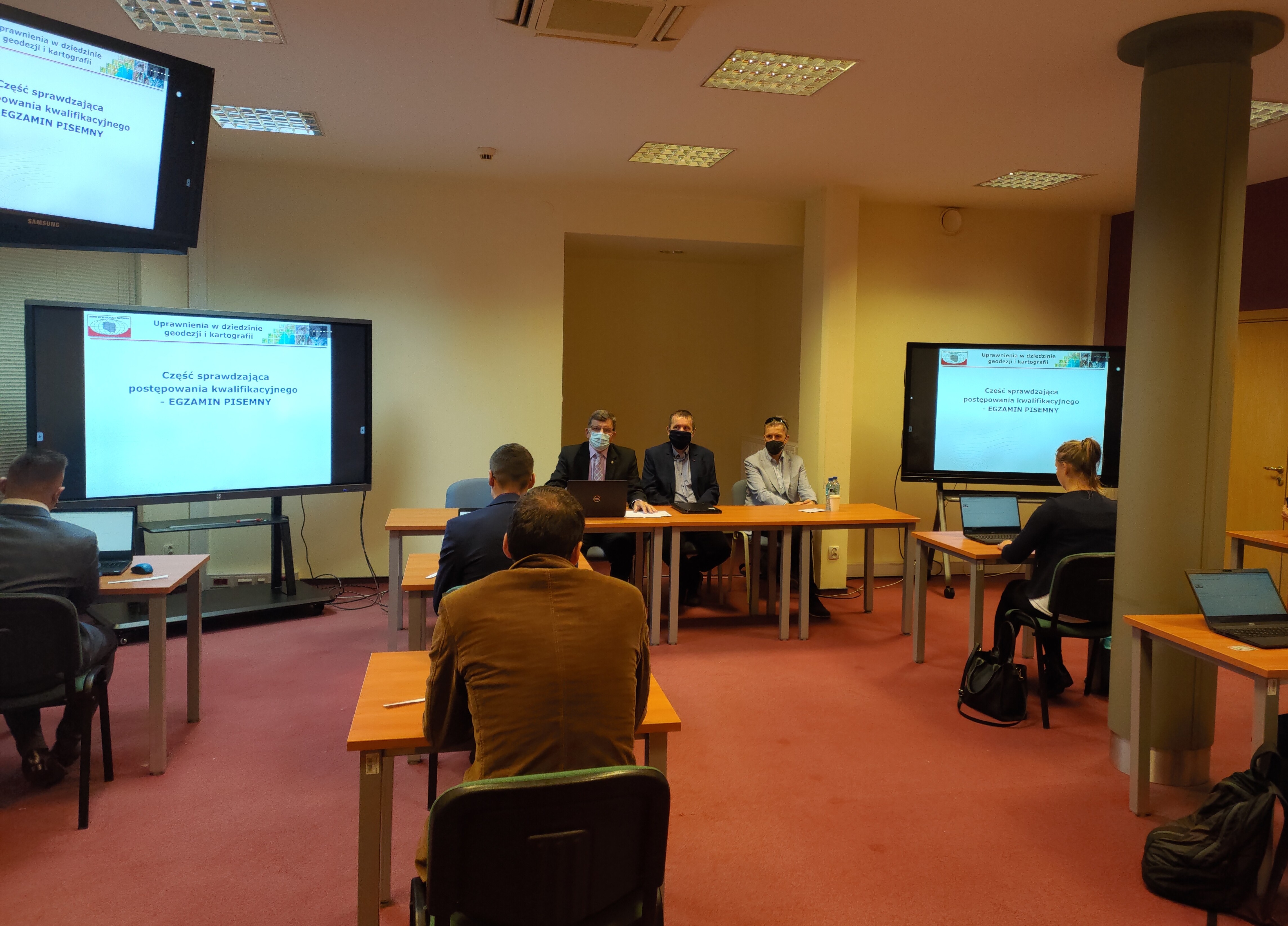 Zdjęcie przedstawia salę egzaminacyjną z z członkami Komisji egzaminacyjnej i osobami przystępującymi do egzaminu siedzącymi pojedynczo przed laptopami.