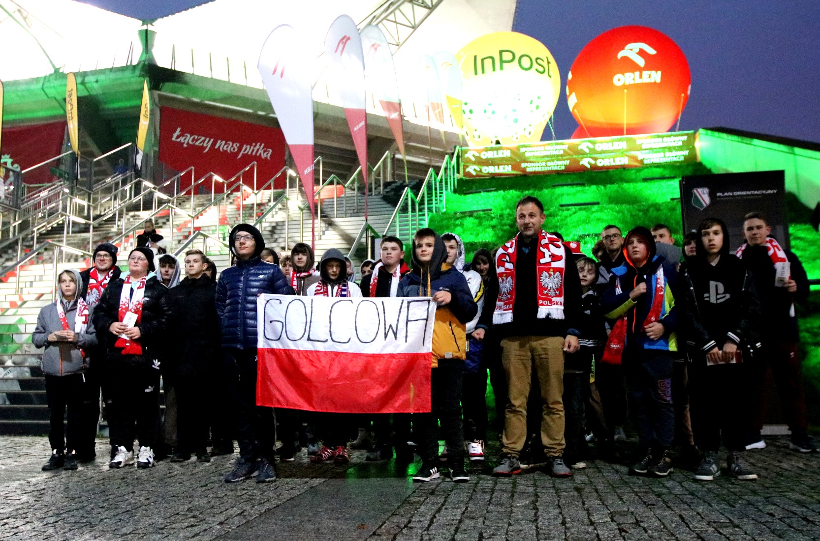 Grupa dzieci i dorosłych stoi pod betonowymi schodami, trzymają biało-czerwoną flagę z napisem Golcowa.