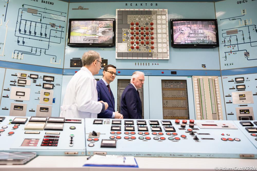 Wnętrze reaktora jądrowego, a w tle premier Mateusz Morawiecki i wicepremier Jarosław Gowin idą i rozmawiają z naukowcem.