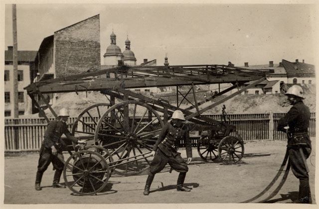 Drabina pożarnicza Zawodowej Pożarnej w Nowym Sączu z 1942 rok a obok niej trzech strażaków. W tle widać zabudowę Nowego Sącza bazyliką Św Małgorzaty.
