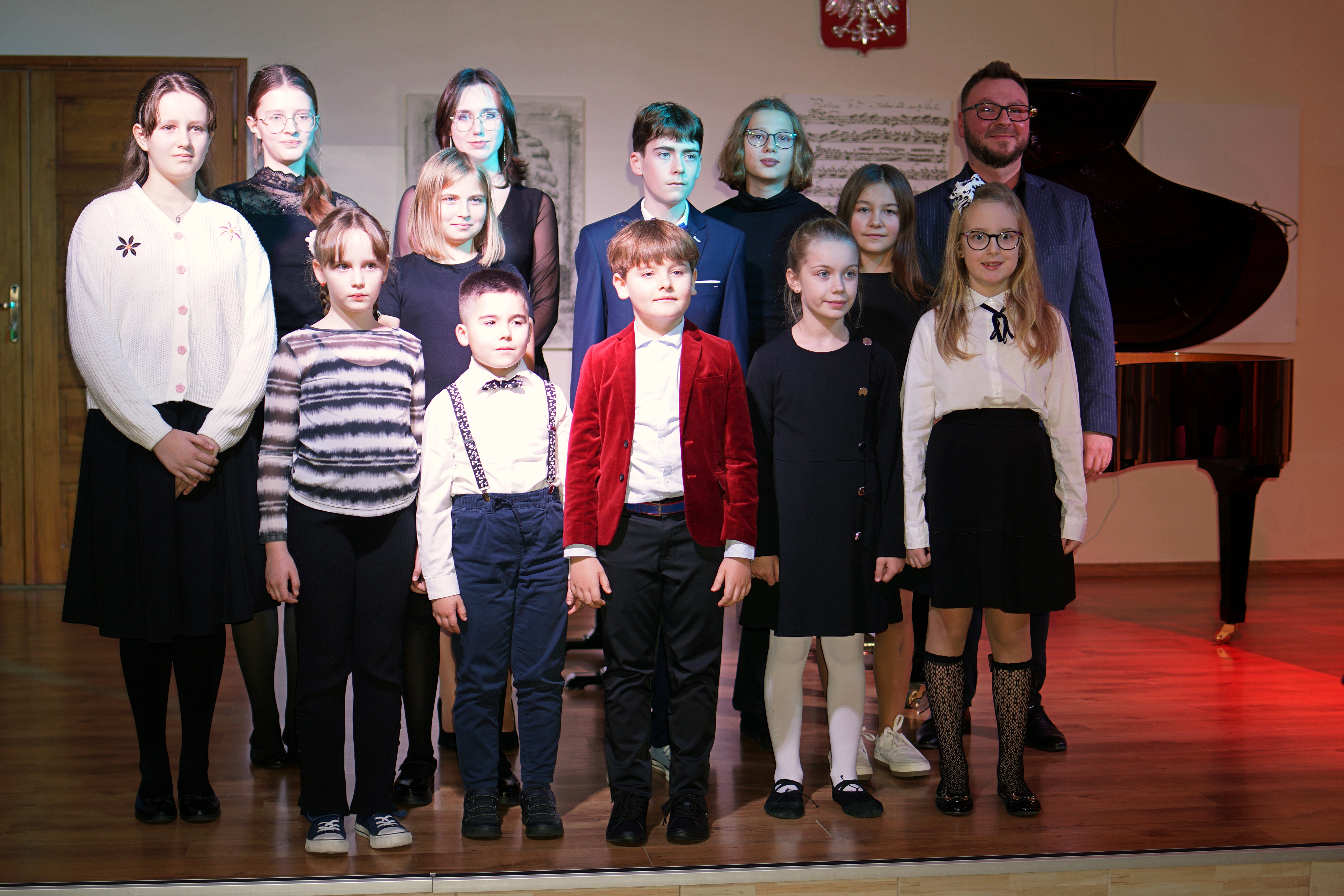 Zdjęcie przestawia dzieci i młodzież stających na scenie Państwowej Szkoły Muzycznej im. J. S. Bacha w Grajewie wraz z nauczycielem. Wszyscy są ubrani na galowo, uśmiechają się i pozują do zdjęcia. W tle widać fortepian, portret J. S. Bacha, grafiki partytury nut, fragment godła Polski oraz drewniane brązowe drzwi. 