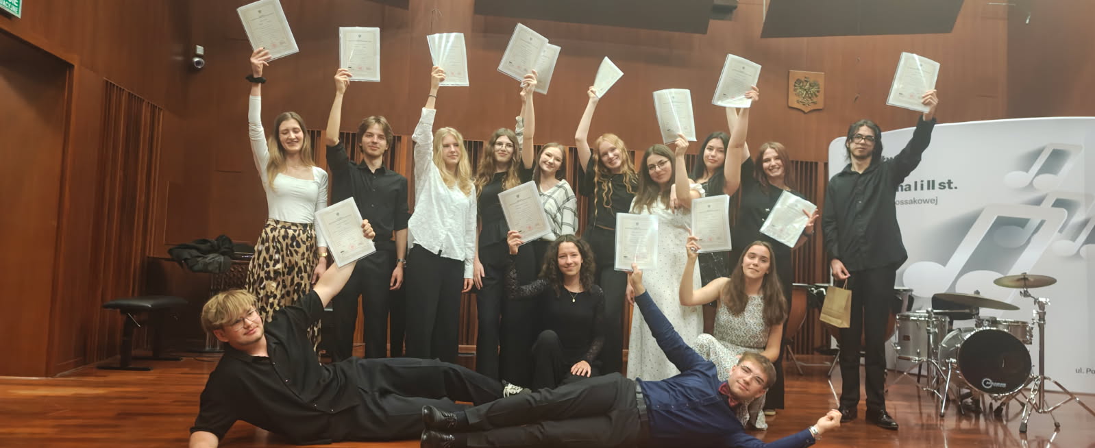 Absolwenci II st. 2026 na scenie w sali koncertowej szkoły. 15 absolwentów z dyplomami w rękach