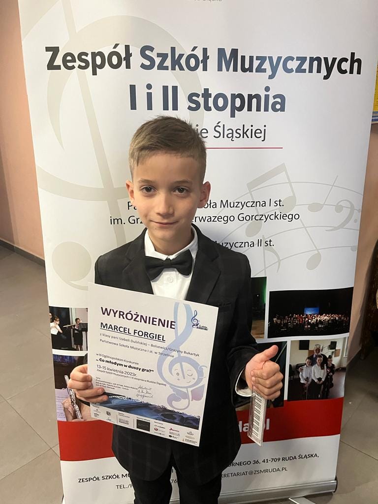 Na zdjęciu widoczny uśmiechnięty uczeń Marcel Forgiel trzymający w rękach dyplom konkursowy. Stoi na tle baneru konkursowego.