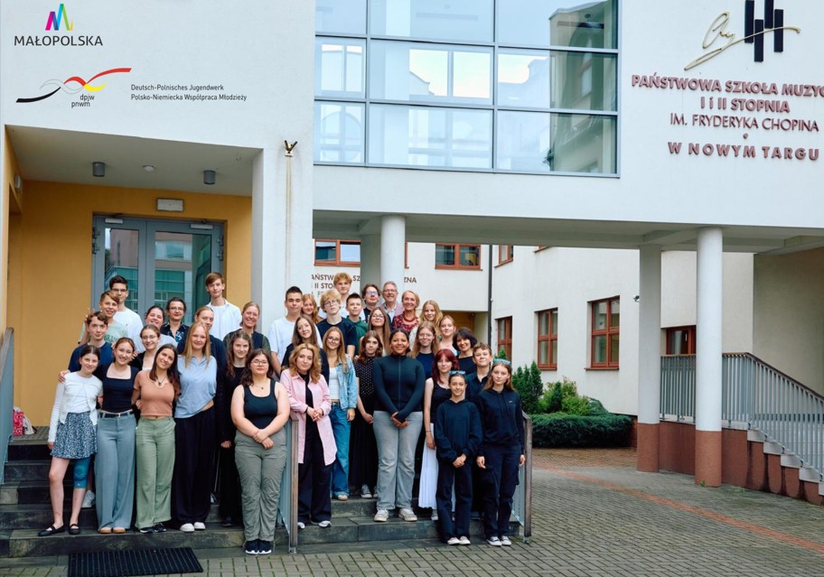 Na zdjęciu widoczna jest duża grupa młodzieży i nauczycieli, którzy stoją wspólnie przed wejściem do budynku szkoły muzycznej. Budynek jest jasny, nowoczesny, z dużymi oknami i przeszklonymi elementami.Na ścianie po prawej stronie widnieje napis: Państwowa Szkoła Muzyczna I i II stopnia im. Fryderyka Chopina w Nowym Targu, obok którego znajduje się ozdobne logo. Po lewej stronie zdjęcia, nad wejściem, znajdują się logotypy: kolorowe logo Małopolski, logo Polsko-Niemieckiej Współpracy Młodzieży (Deutsch-Polnisches Jugendwerk). Grupa, licząca około 35 osób, stoi na schodach i przed wejściem, ustawiona w kilku rzędach. Widać uśmiechnięte twarze, część osób obejmuje się ramionami, co podkreśla przyjazną i koleżeńską atmosferę. Zdjęcie ma charakter pamiątkowy i oficjalny, prawdopodobnie wykonane zostało przy okazji projektu wymiany młodzieży lub wydarzenia artystyczno-edukacyjnego.