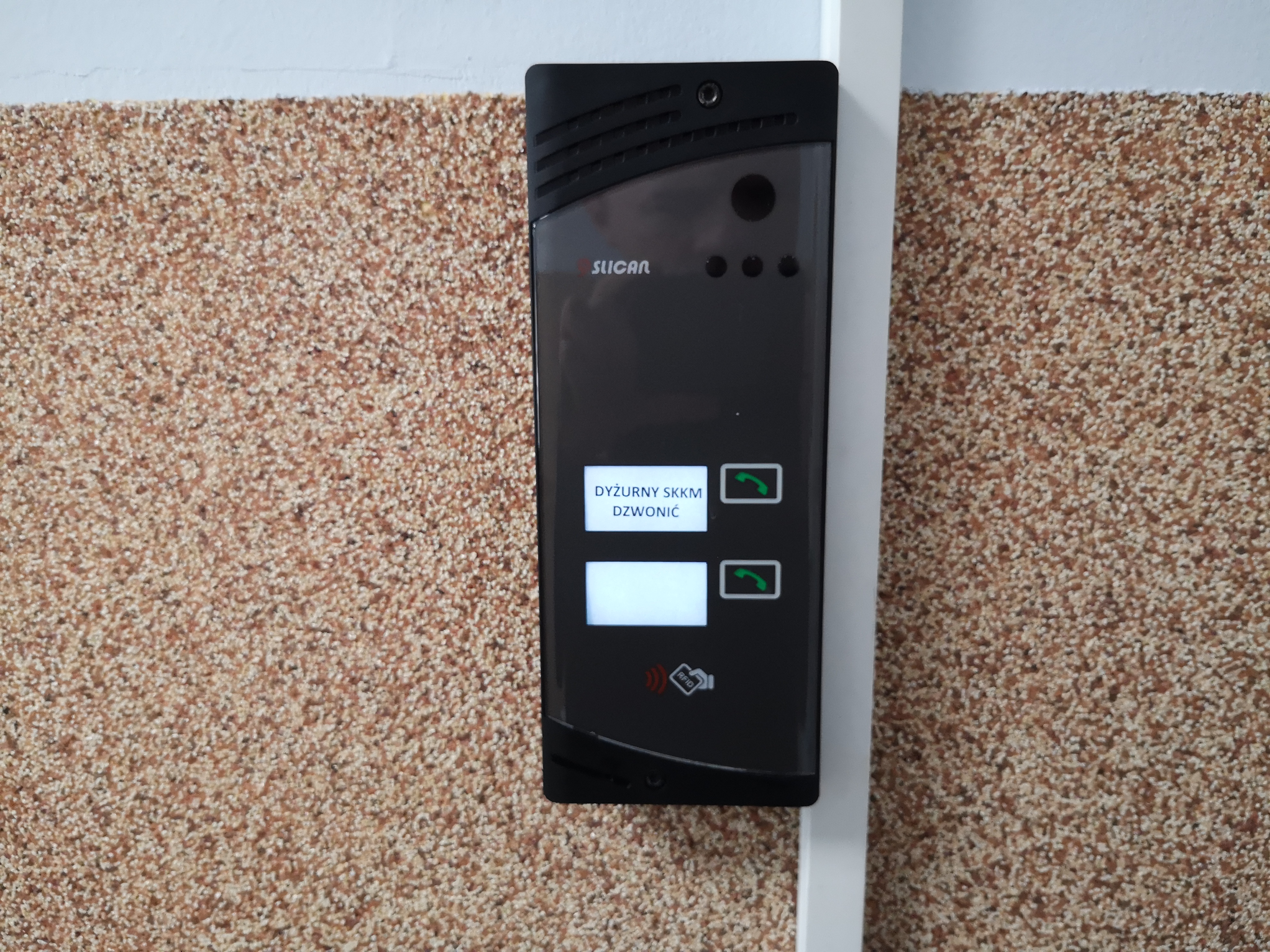 Zdjęcie przedstawia interkom. Interkom znajduje się w centralnej części zdjęcia. Interkom jest koloru czarnego. Znajduje się na ścianie o dwóch kolorach - szarej w górnej części oraz brązowej w dolnej części. Interkom ma dwa przyciski z symbolem zielonej słuchawki. Obok nich znajdują się białe pola. Górny przycisk uruchamia rozmowę z dyżurnym - wskazuje to napis na białym polu przy przycisku o treści Dyżurny SKKM dzwonić.