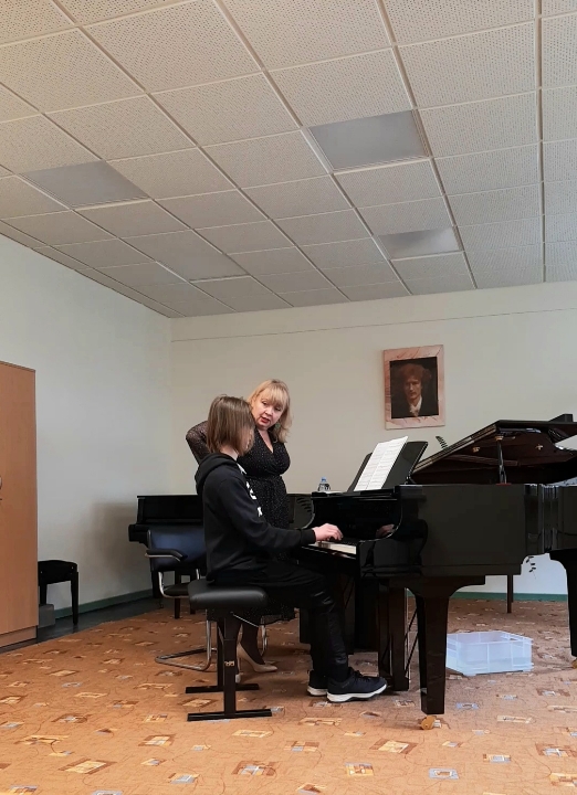 Uczeń siedzący przy fortepianie, obok niego nauczycielka. Po lewej stronie widoczna szafa. Na ścianie wisi portret Ignacego Jana Paderewskiego.