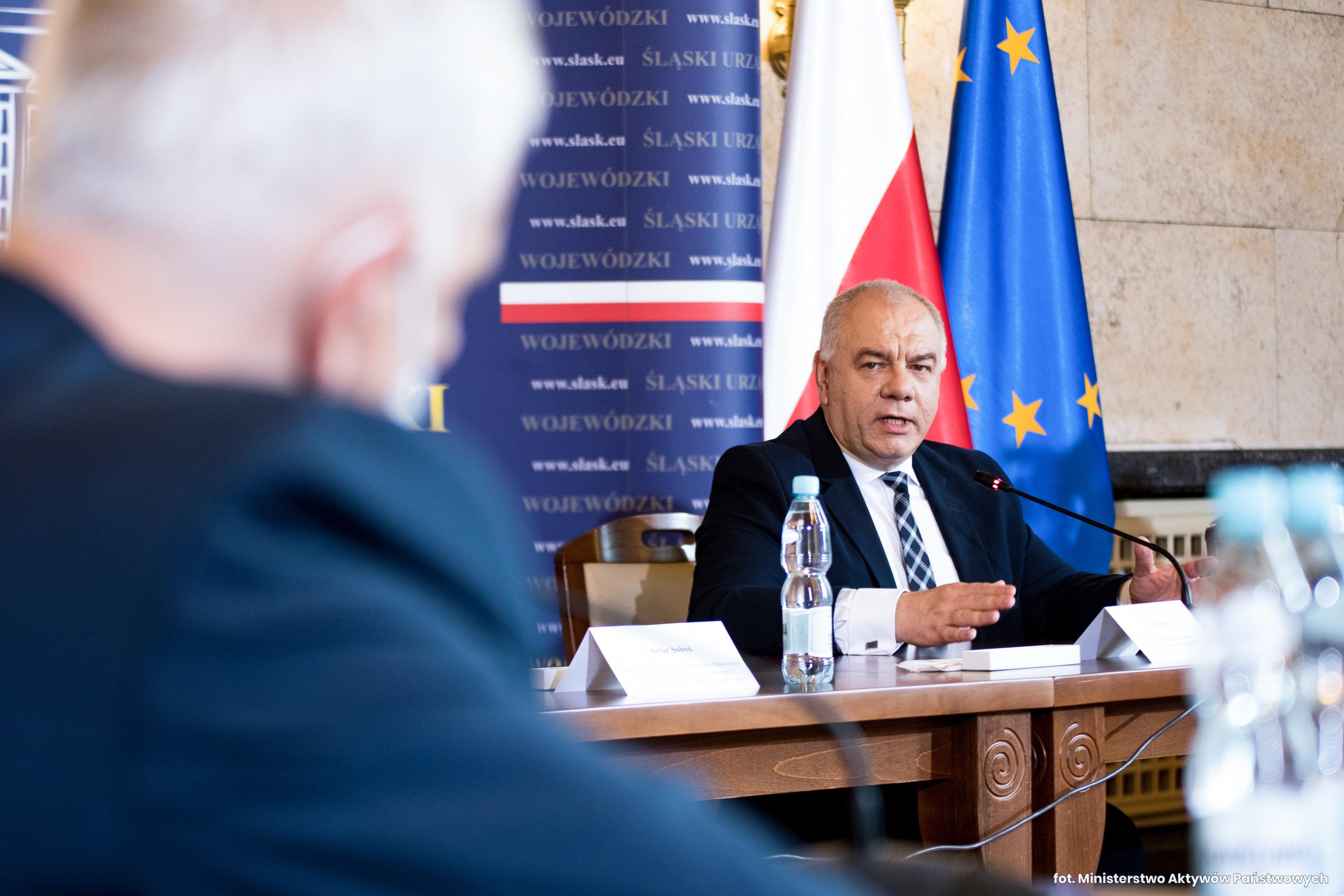 Wicepremier Jacek Sasin podczas spotkania w Katowicach. Wicepremier siedzi za stołem prezydialnym. Przed nim Dominik Kolorz. Wicepremier przemawia.