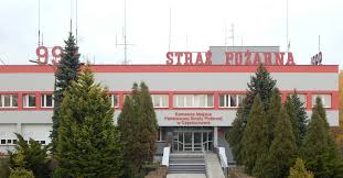 Zdjęcie frontu budynku Komendy Miejskiej Państwowej Straży Pożarnej w Częstochowie wraz z wejściem głównym. Elewacja budynku w kolorze szarym z czerwonymi pasami w linii okien oraz dachu. Na dachu duży napis "998 STRAŻ POŻARNA)