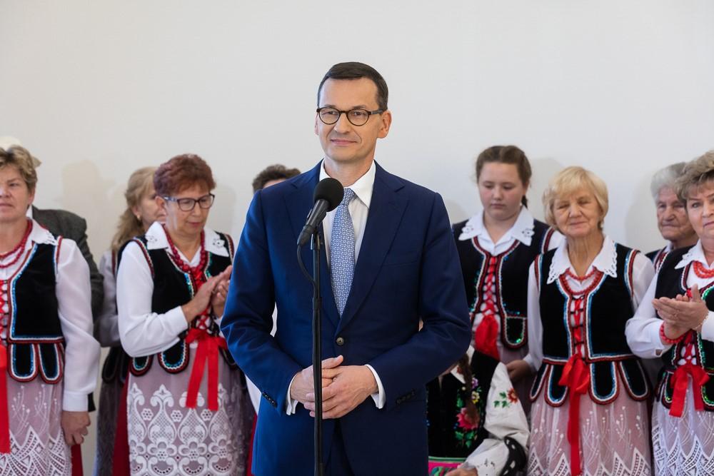 Premier Mateusz Morawiecki stoi na tle koła gospodyń wiejskich.