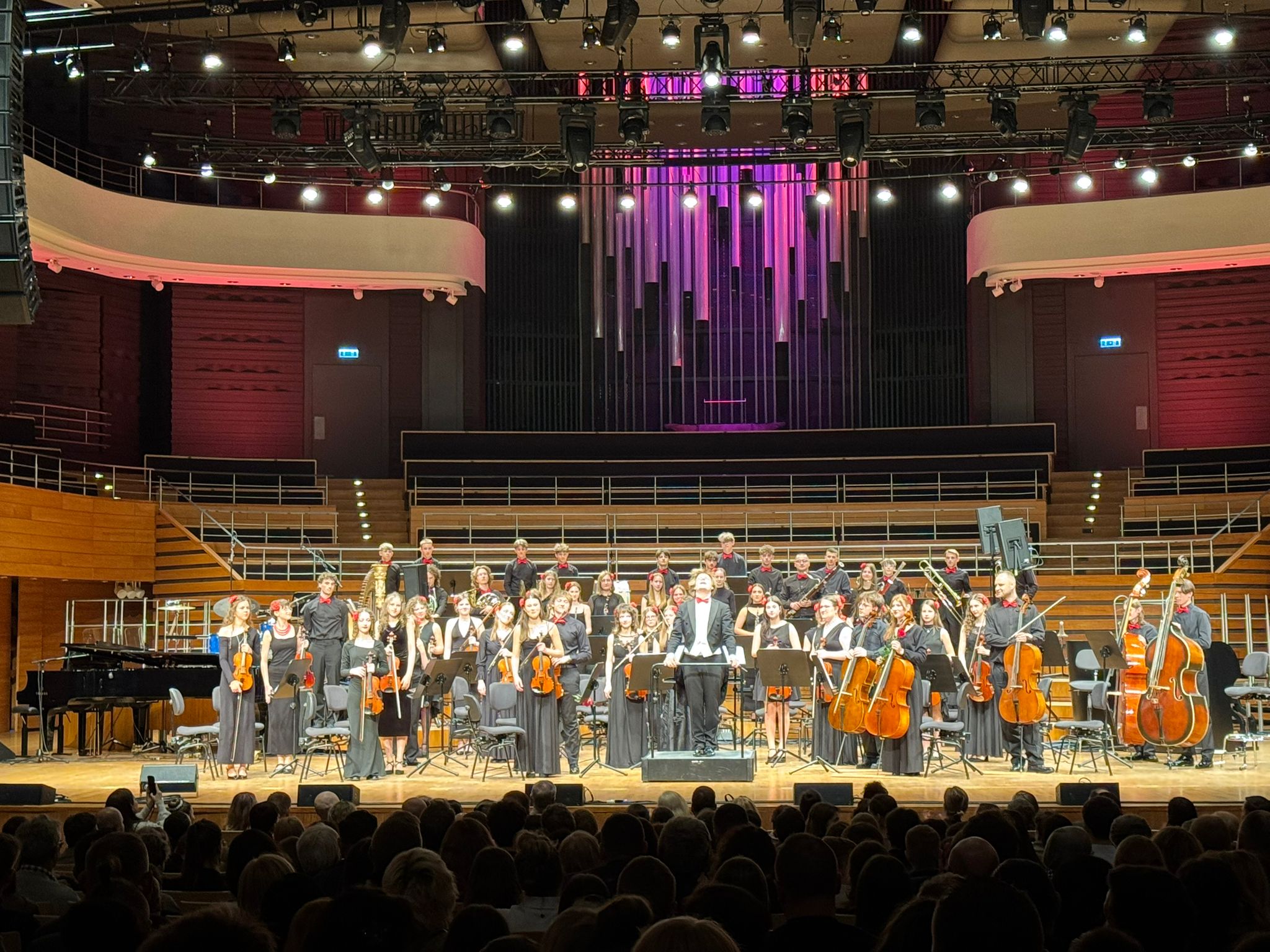 Zdjęcie zrobione z perspektywy widowni, przedstawia młodzieżową orkiestrę symfoniczną PSM II st. im. Stanisława Moniuszki w Jeleniej Górze na scenie Narodowego Forum Muzyki we Wrocławiu. Muzycy, ubrani w eleganckie czarne stroje z czerwonymi muchami, artystki z kwiatami przypiętymi we włosach, stoją wzdłuż krawędzi sceny, trzymając swoje instrumenty, m.in. skrzypce, wiolonczele i kontrabasy. W centrum, na podwyższeniu, stoi dyrygent w czarnym fraku i czerwonej muszce, z batutą skierowaną w stronę orkiestry. Scena jest oświetlona jasnym światłem reflektorów, a w tle widać imponujące organy piszczałkowe z fioletowym i różowym podświetleniem. Po lewej stronie sceny stoi czarny fortepian, a po prawej – zestaw perkusyjny. Całość tworzy uroczystą i podniosłą atmosferę, podkreśloną przez eleganckie wnętrze sali koncertowej.