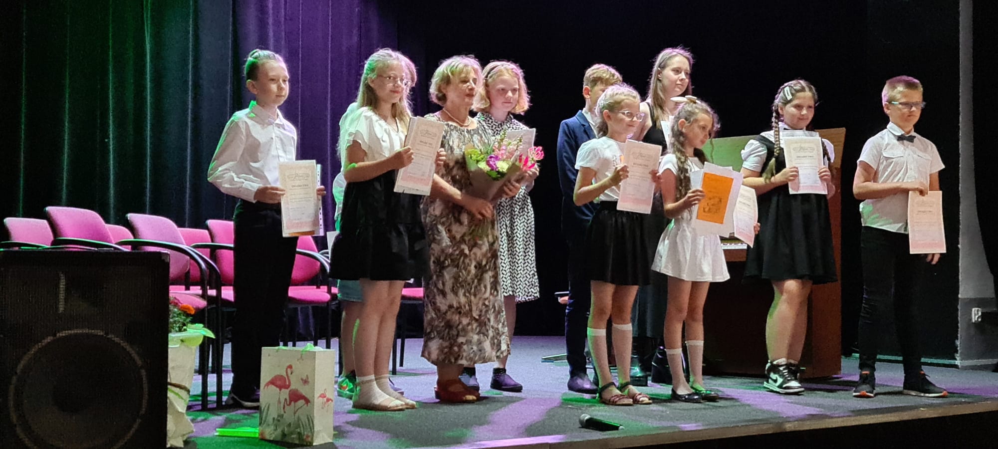 Grupa uczniów trzymająca dyplomy wraz z nauczycielem p. Martą Dąbrowską stoją na scenie w sali koncertowej w Szczytnej