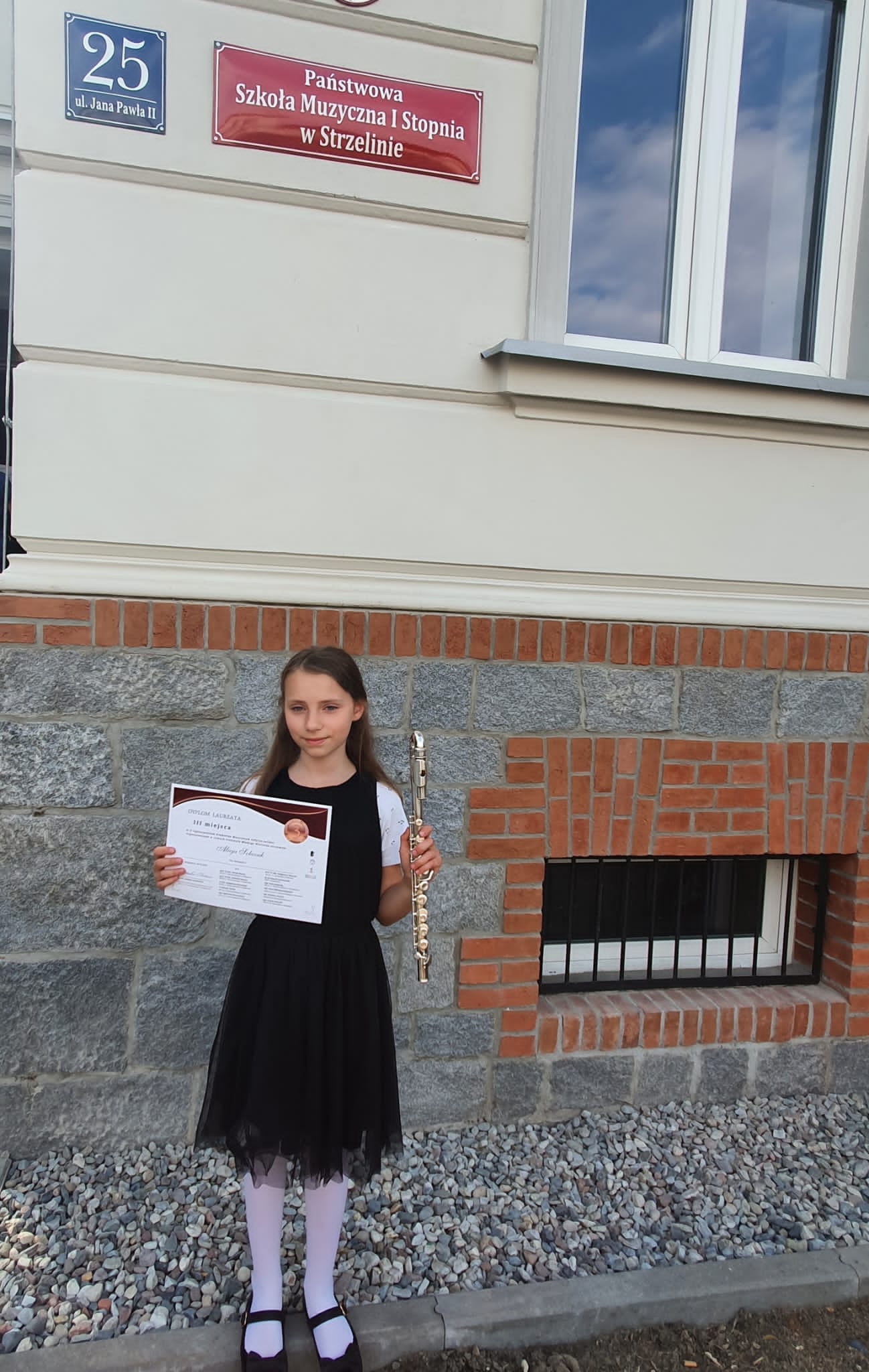 Na zdjęciu znajduje się elegancko ubrana dziewczynka, trzymająca w dłoniach flet oraz dyplom. Stoi na tle budynku PSM Strzelin.