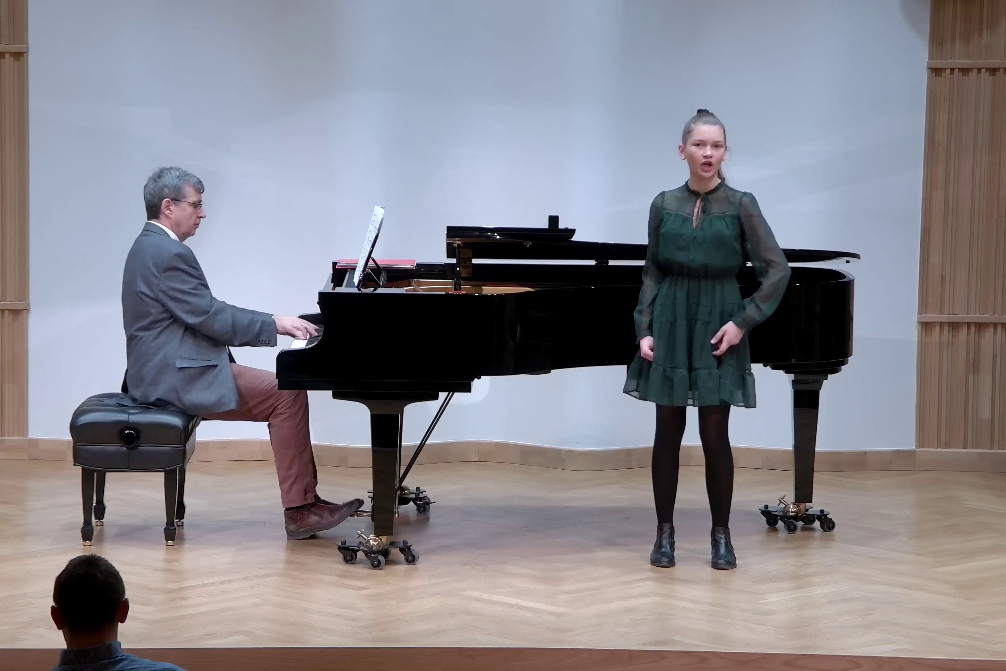 Zdjęcie. Na estradzie występuje ubrana w zieloną sukienkę śpiewaczka z towarzyszeniem pianisty, który gra na dużym czarnym fortepianie.