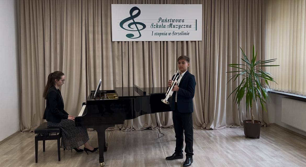 Na zdjęciu widoczny jest trzymający w rękach trąbkę uczeń oraz siedzący przy fortepianie akompaniator.