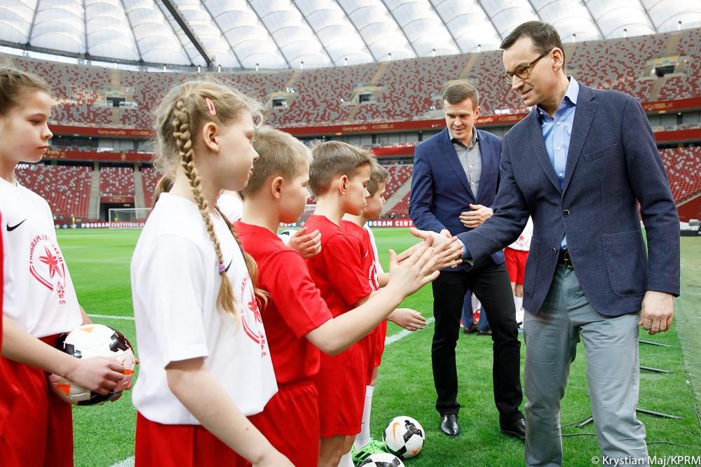 Premier Mateusz Morawiecki przybija piątkę z dziećmi ze szkółki piłkarskiej.