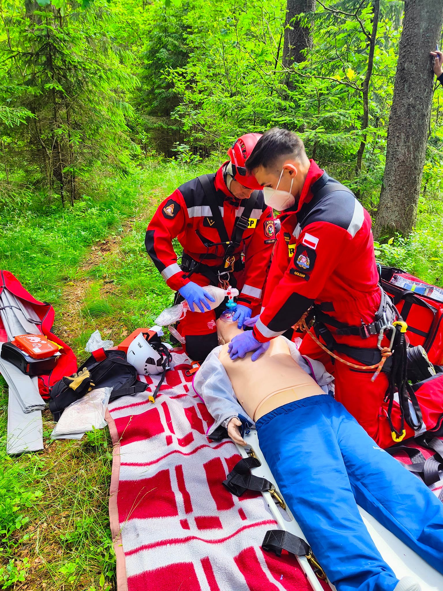 Dwóch strażaków - ratowników medycznych wykonuje resuscytację krążeniowo - oddechową u poszkodowanego (manekin w ćwiczeniach), wokół teren lesny