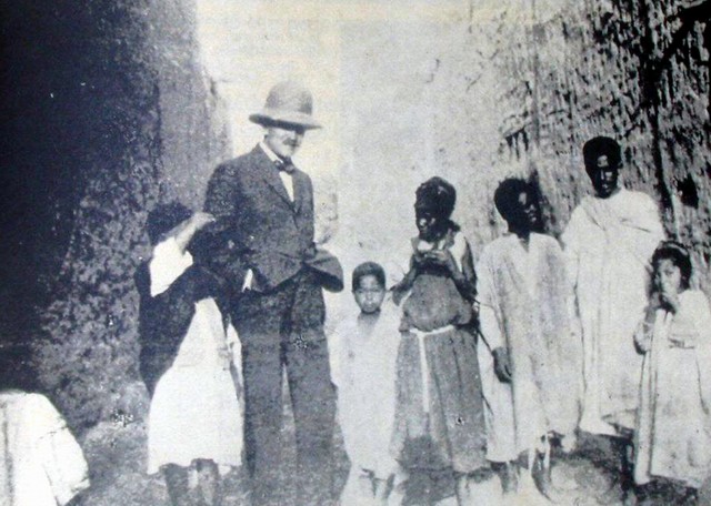Czarno-białe zdjęcie z Tunisu 1914 r. Na zdjęciu Szymanowski stoi w grupie ciemnoskórych miejscowych dorosłych i dzieci. Po obu stronach kamienne ściany. Tubylcy w luźnych, długich, płóciennych strojach. Szymanowski w marynarce i białej koszuli z muszką. Na głowie korkowy kapelusz, kojarzący się z wyprawami do Afryki.