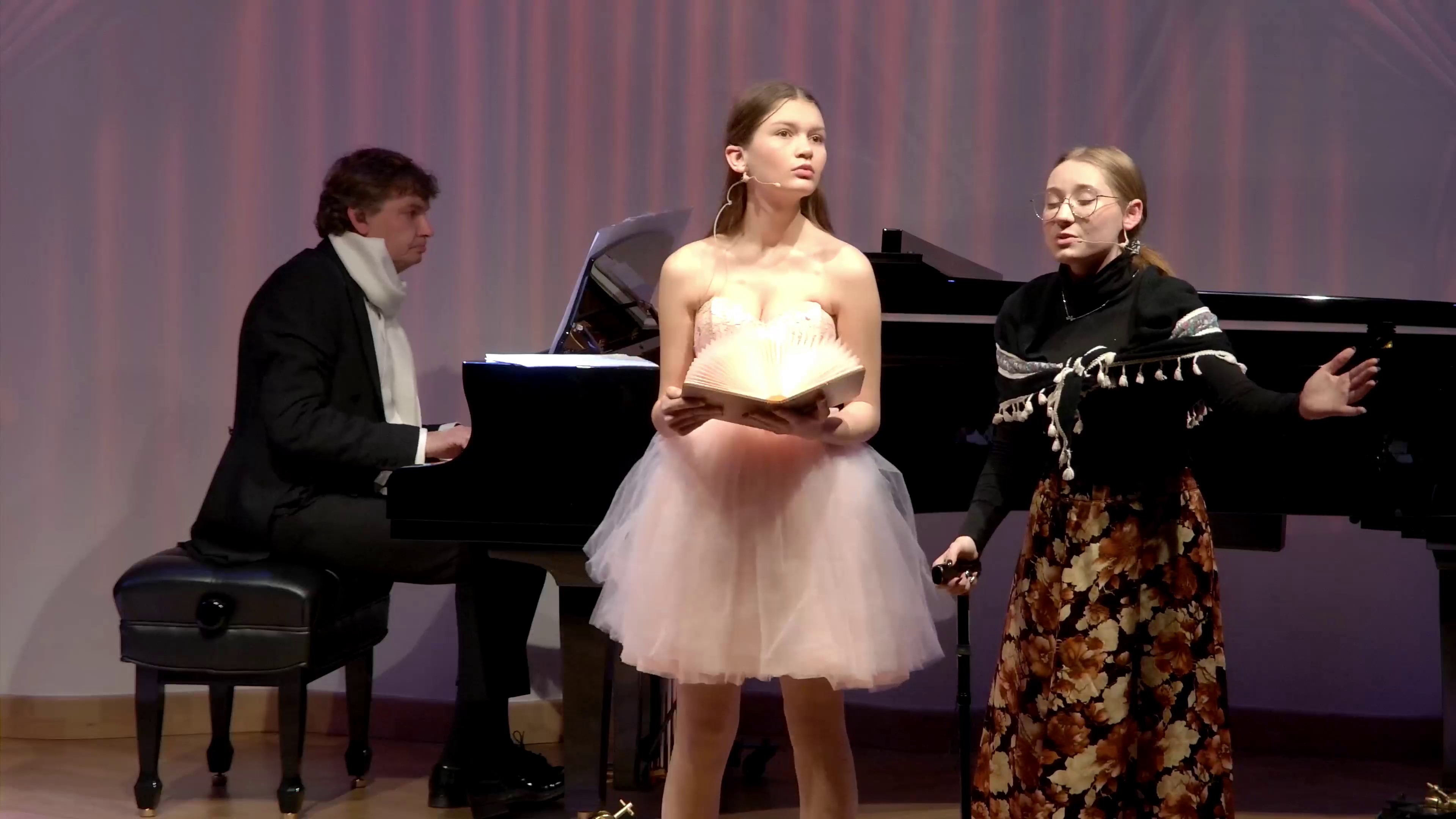 Zdjęcie. Na estradzie sali koncertowej występują dwie młode śpiewaczki. Towarzyszy im przy fortepianie pianista grający na dużym czarnym fortepianie.