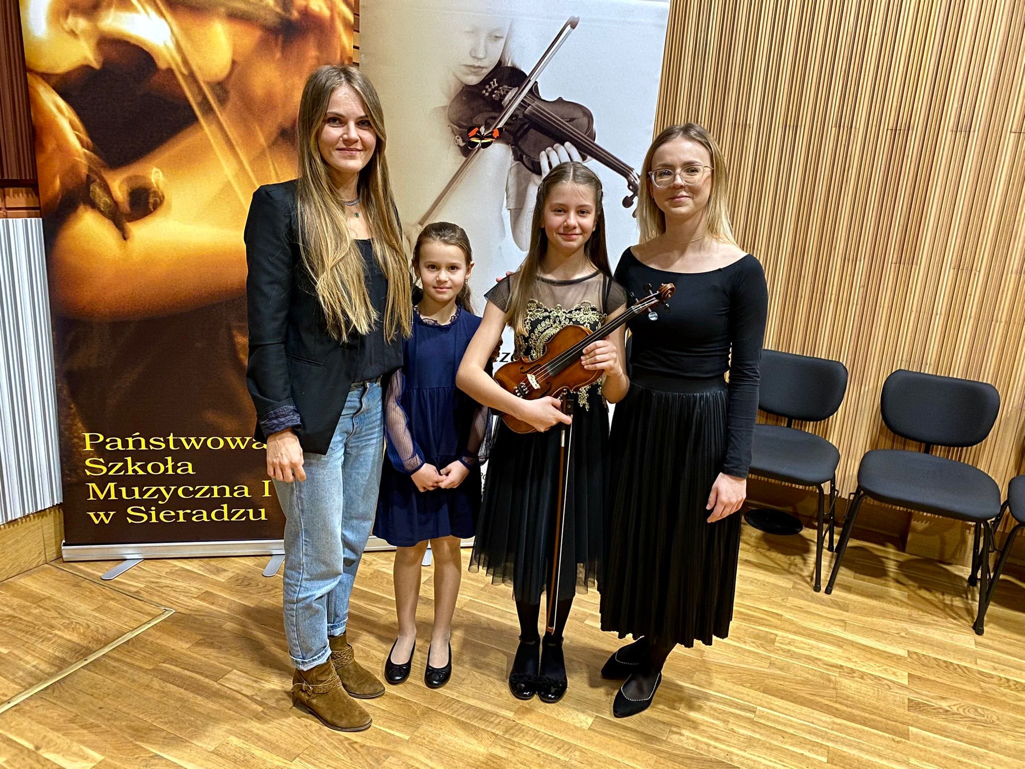 Na fotografii znajduje się nauczycielka Edyta Kornafel, uczennice Zuzanna Szczepańska oraz trzymająca w rękach skrzypce Agata Kostanowicz oraz nauczycielka Monika Duda. W tle widoczne są dwa duże szkolne roll-upy. 