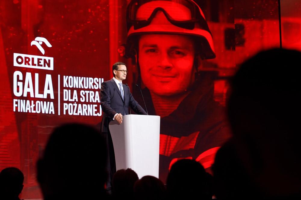 Premier Mateusz Morawiecki, a w tle ścianka wizyjna ze strażakiem.